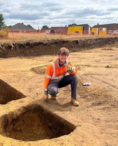 A man crouches in front of an excavation, giving a thumbs up. 